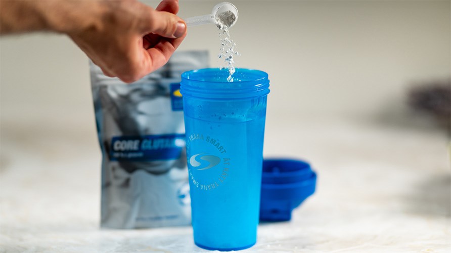 Käsi kaataa mitallista Core Glutamine -jauhetta siniseen sheikkeriin. Käsi kaataa mitallista Core Glutamine -jauhetta siniseen sheikkeriin.